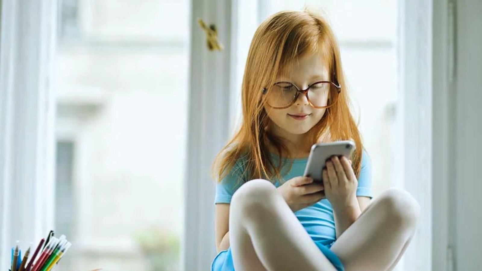 Young girl with red hair and glasses sitting by a window, looking at a smartphone while holding it with both hands.
