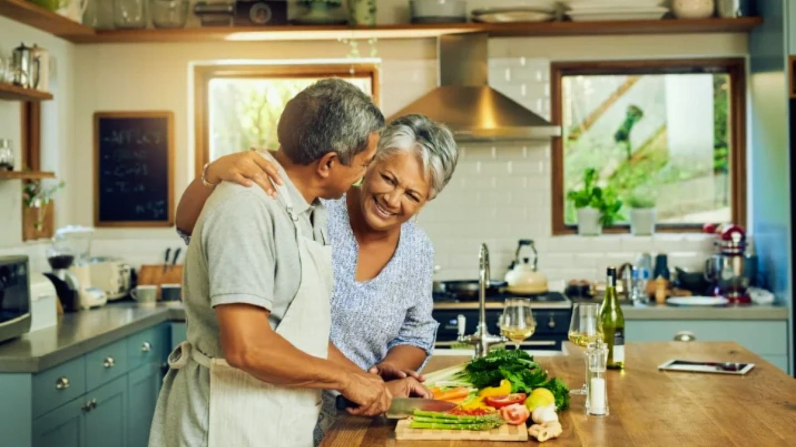 An older couple in a kitchen, happily cutting vegetables together on a wooden countertop.