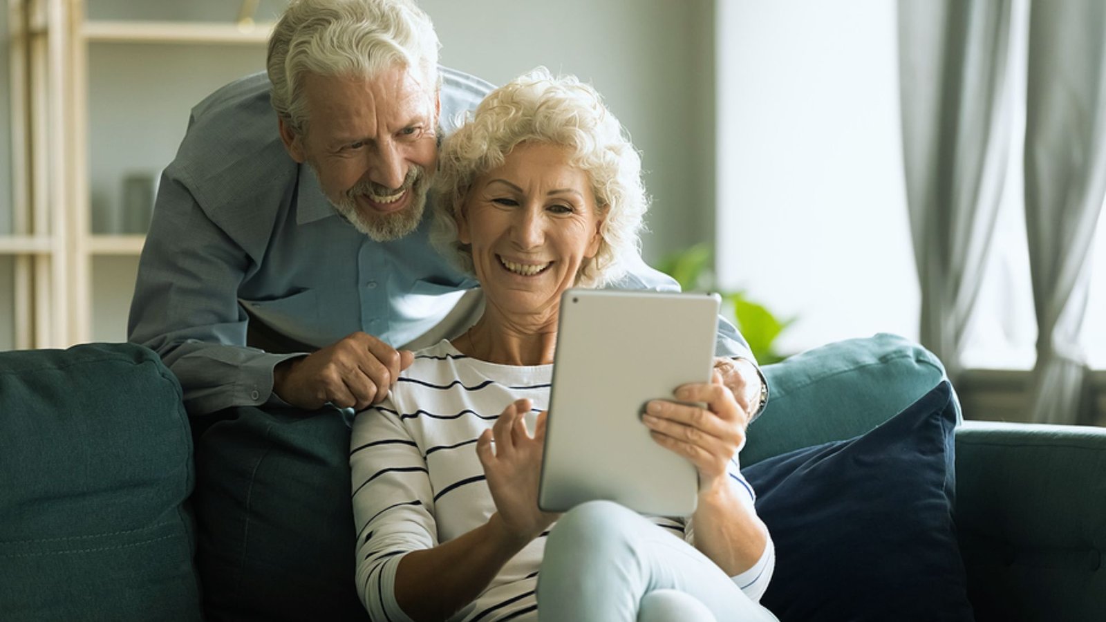 An older couple sits on a couch, smiling as they look at a tablet together.