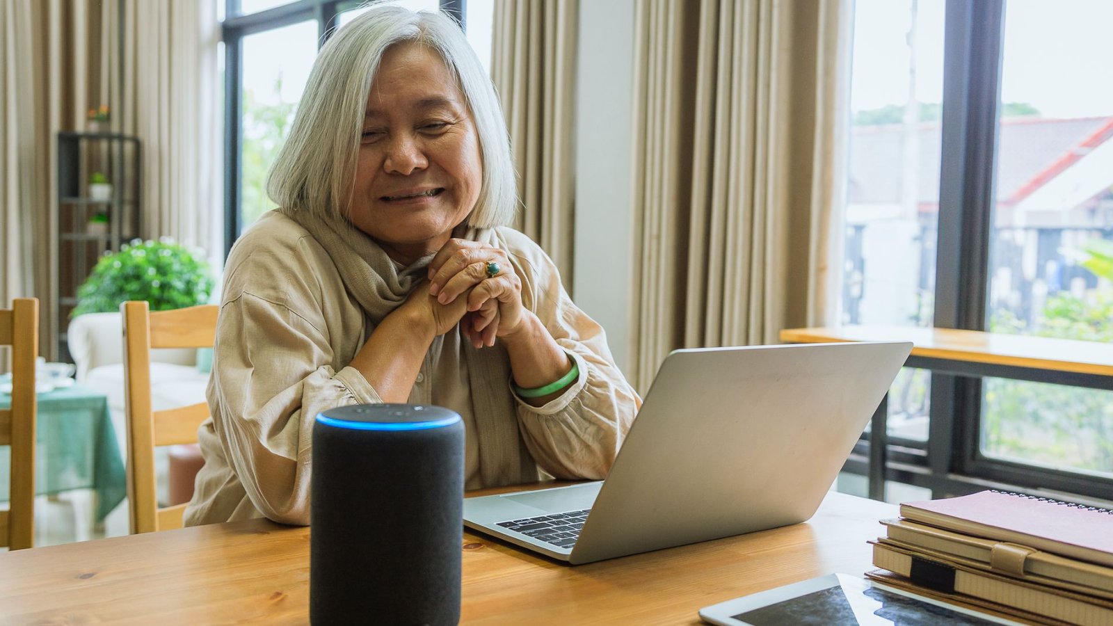  An older woman sits at a table with a laptop and an Amazon Echo, engaged in a conversation or task.