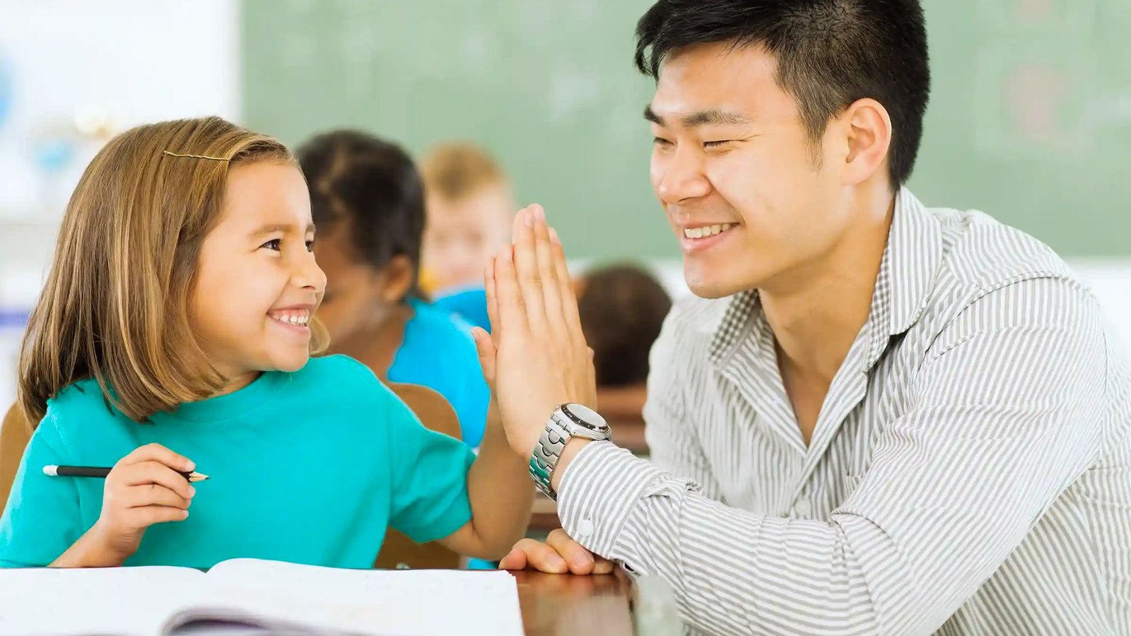 Smiling teacher and young student giving each other a high five in a classroom while the child holds a pencil and sits at a desk.
