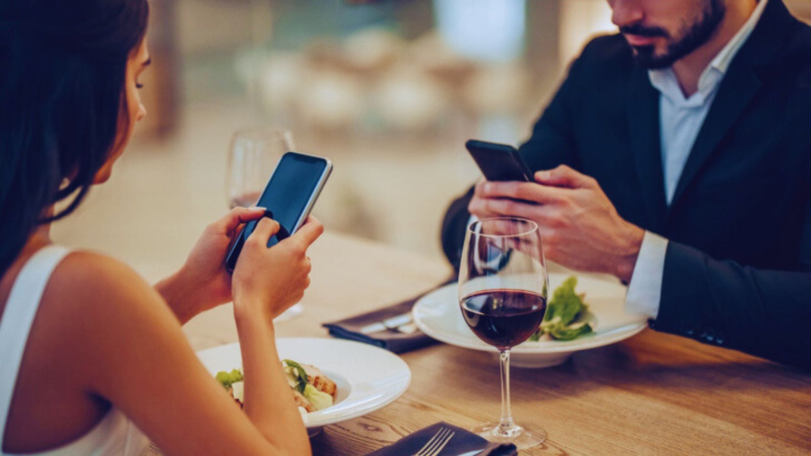  A man and woman sit at a table, each focused on their smartphones, with a casual atmosphere around them.