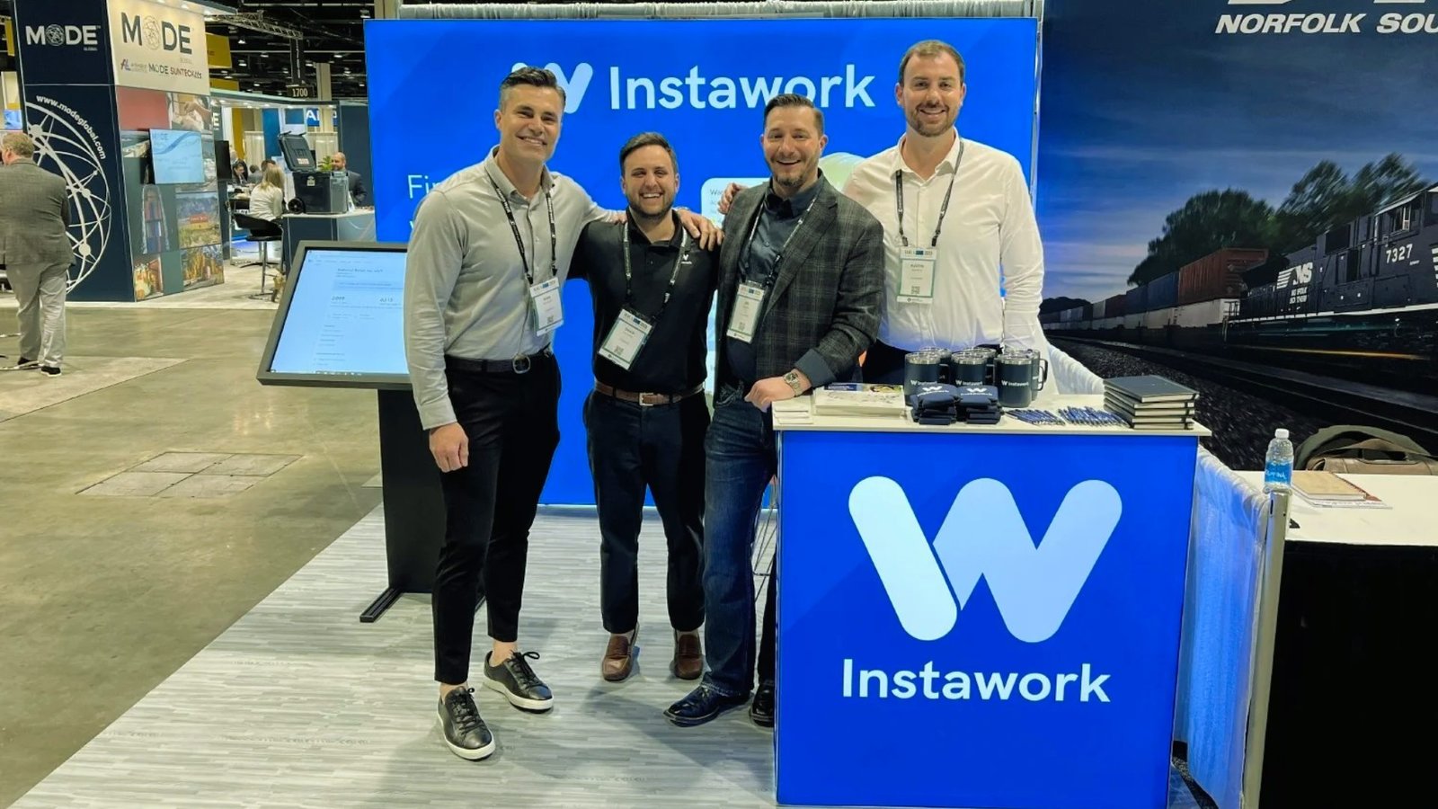  Three men stand in front of a booth at an event, engaging with attendees and showcasing products or services.