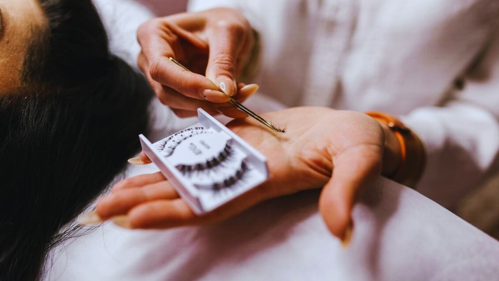A woman sits in a salon chair while a technician applies eyelash extensions to her lashes.