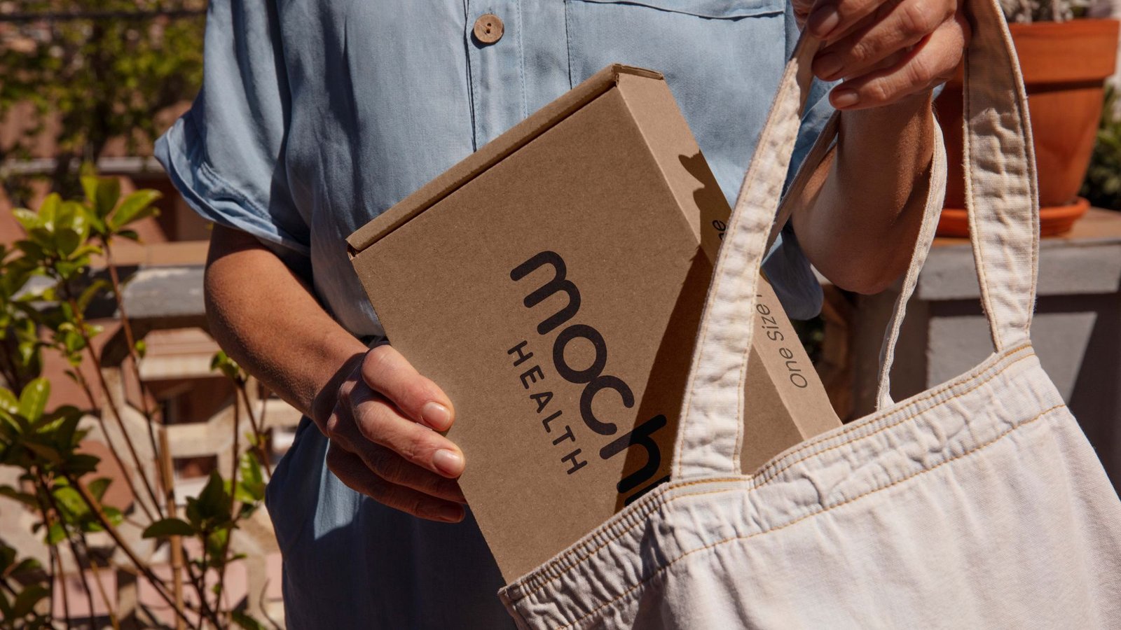 A woman holds a box in one hand and a bag in the other, standing against a neutral background.
