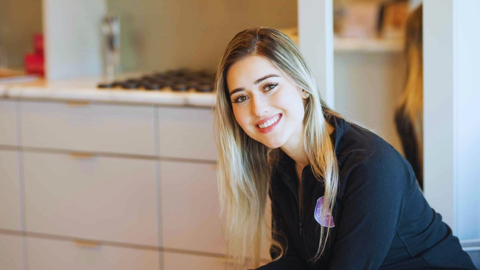 A woman smiling while sitting on a kitchen counter, surrounded by kitchen items and warm lighting.