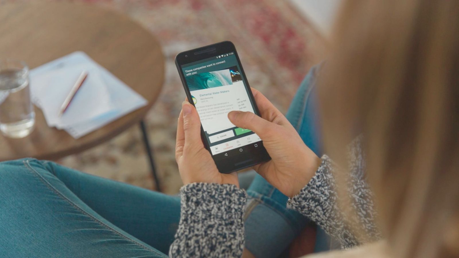 Person sitting indoors and using a smartphone with both hands, viewing an app screen while a table with paper, pencil, and a glass of water sits nearby.
