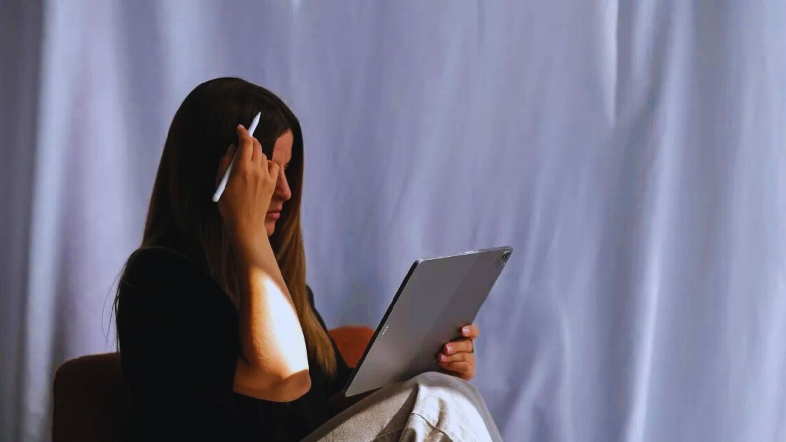 A woman sits on a chair, head in hands, looking stressed while working on a laptop.