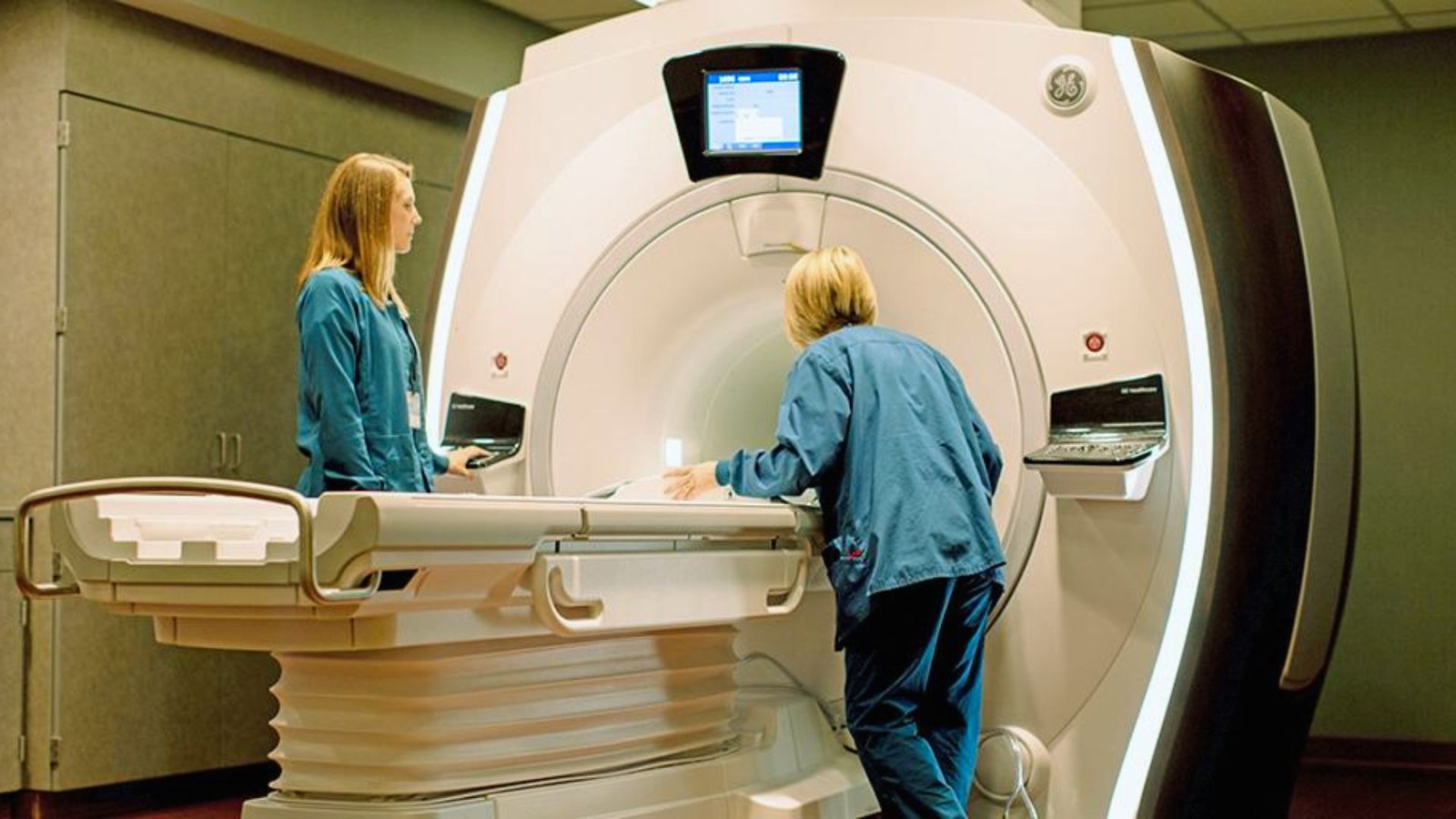 Two women stand beside an MRI machine in a medical setting, discussing the equipment and its purpose.