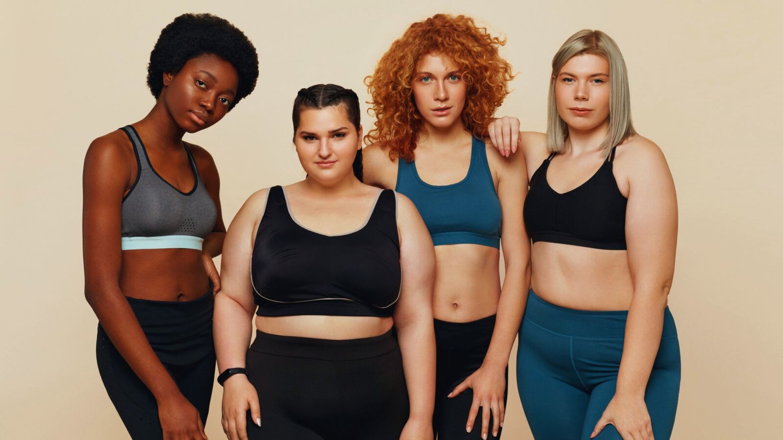 Four women in sports bras smiling and posing together for a group photo in a bright outdoor setting.