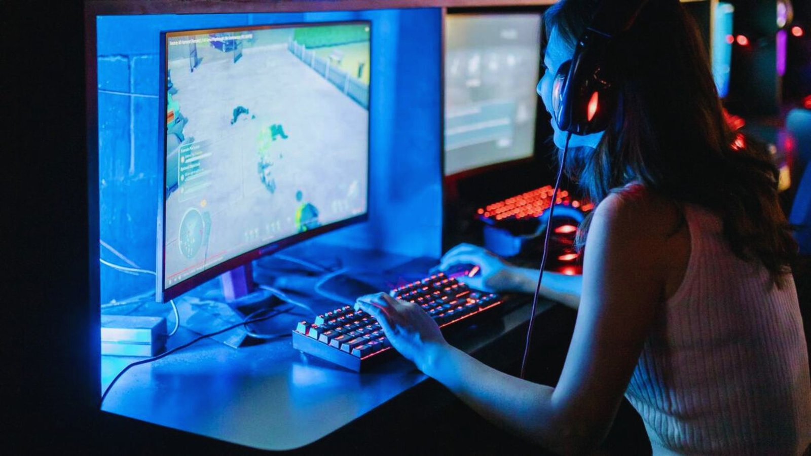  A woman focused on playing video games on her computer, with a gaming headset and colorful LED lights in the background.