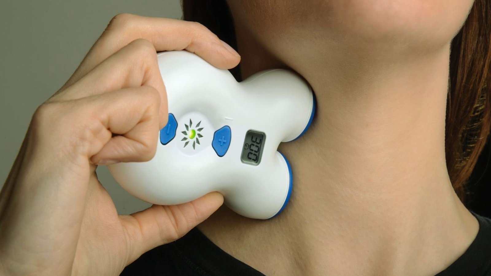 A woman holds an electronic device against her neck, appearing focused and engaged with the technology.