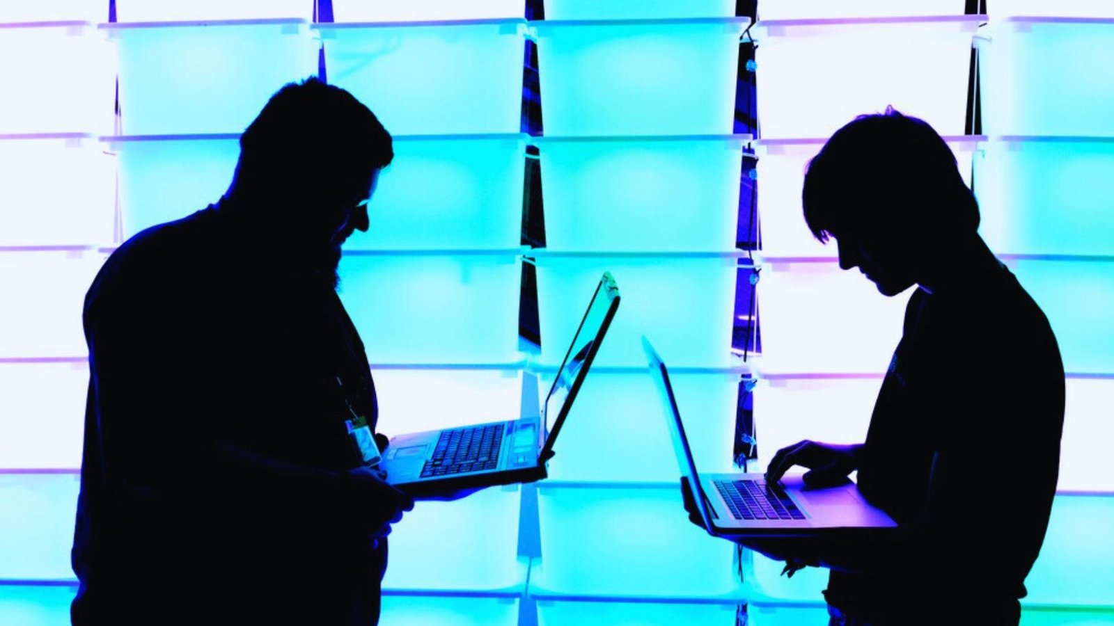  Two silhouetted individuals sit against a wall, each using a laptop, creating a focused and collaborative atmosphere.