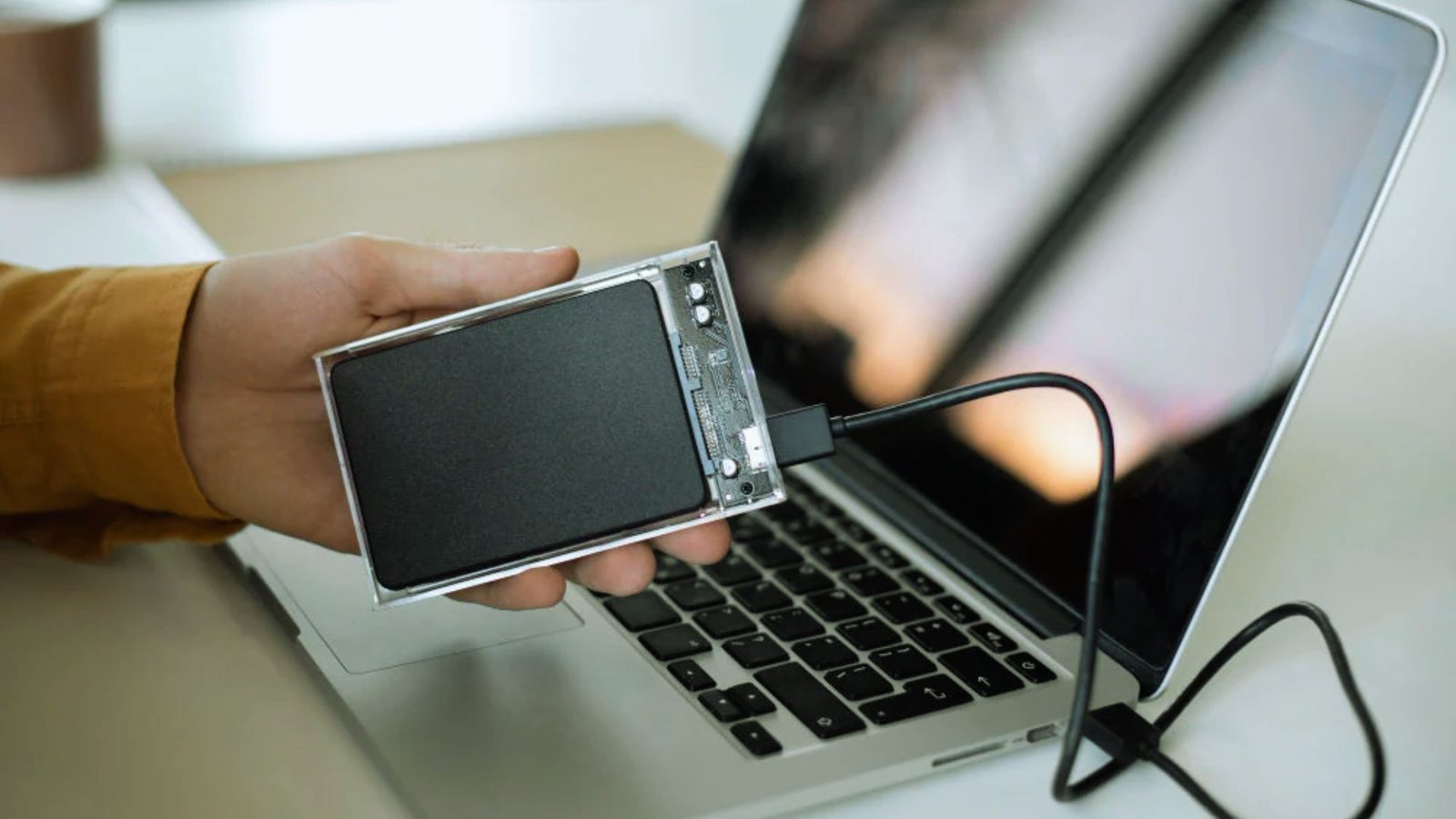 A person holds a portable hard drive in front of an open laptop, indicating data transfer or backup activity.