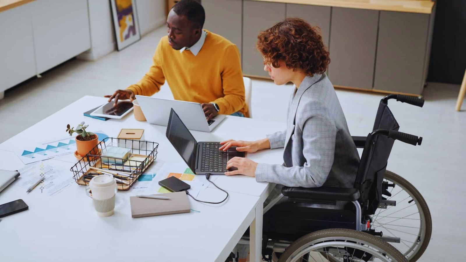  A man in a wheelchair sits at a table with a woman working on a laptop, engaged in conversation.