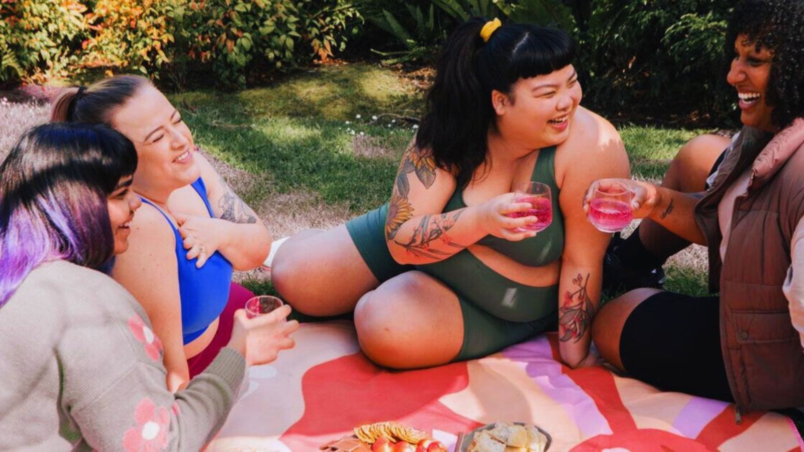 Four women sitting on a blanket outdoors, enjoying drinks and sharing laughter in a relaxed setting.