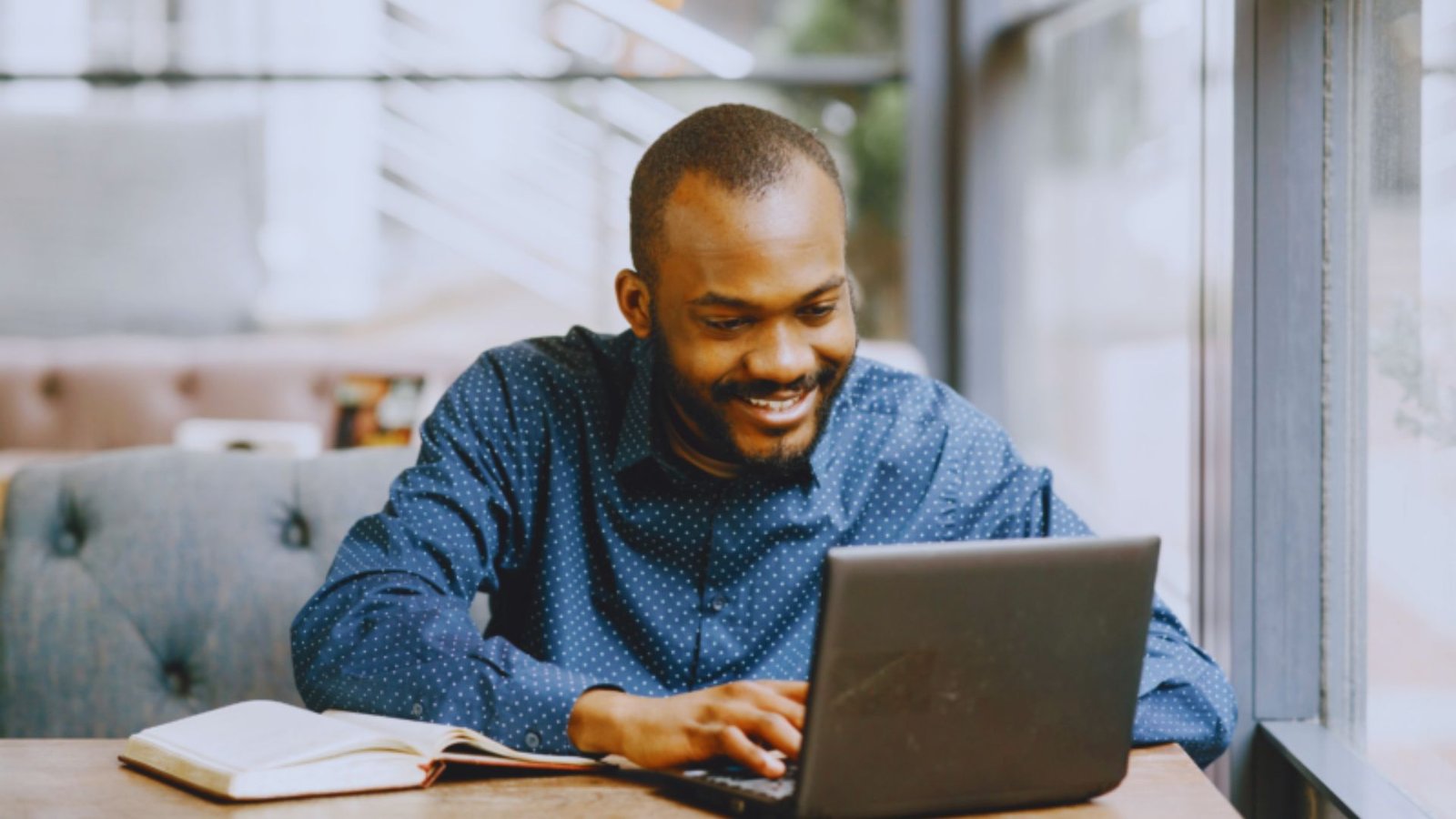  A smiling man sits at a table, working on a laptop, creating a welcoming and productive atmosphere.