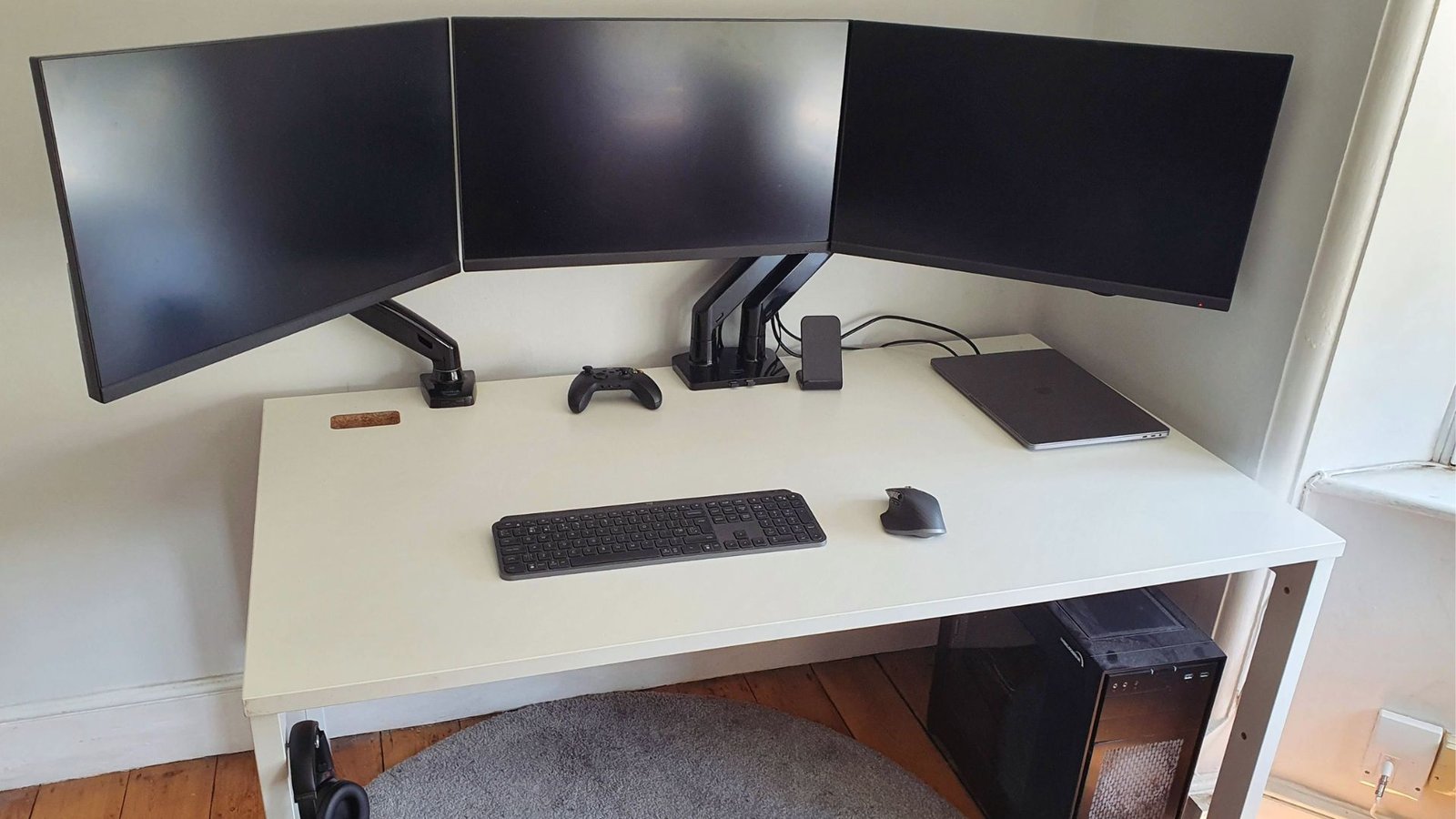 A sleek, modern home office setup with three curved monitors on a white desk. Includes a keyboard, mouse, game controller, and a closed laptop.