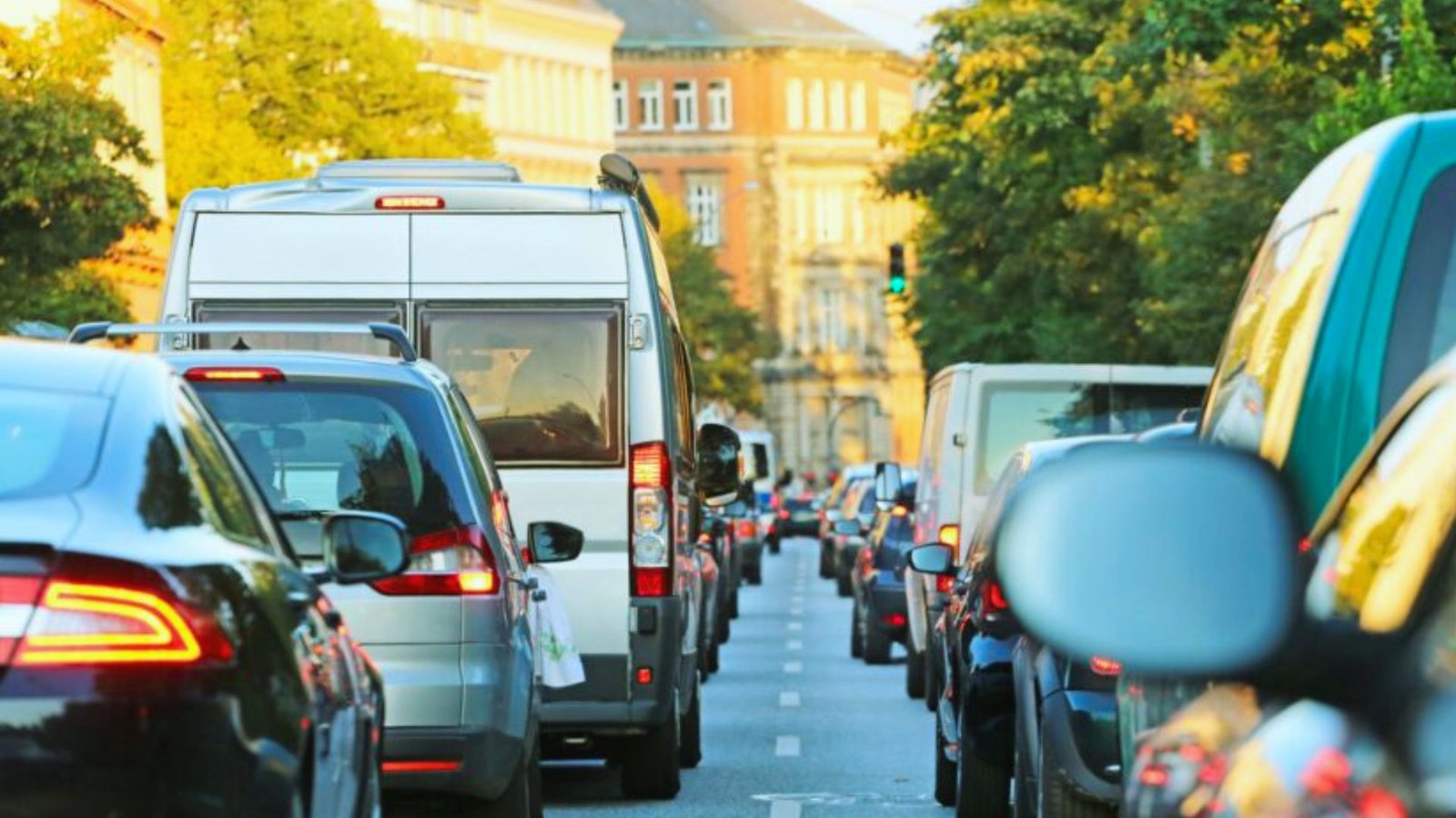 Heavy traffic on a city street, with cars and vans lined up closely. Sunlight reflects off vehicles, while trees and a historic building frame the scene.