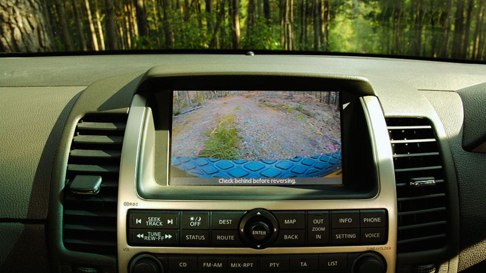Car dashboard with a backup camera display showing a gravel path lined with trees. The screen advises to “Check behind before reversing.”